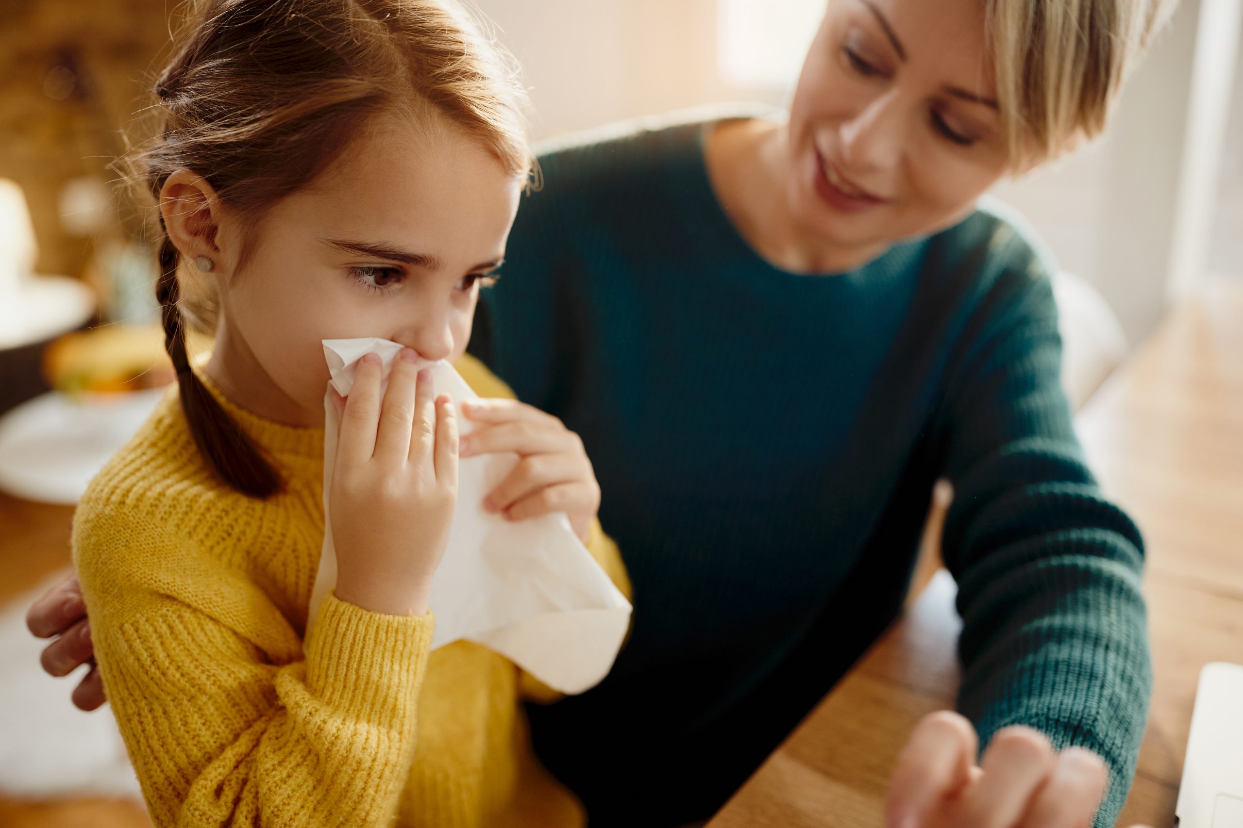 Young girl blowing her nose with a tissue while sitting at home with her mother.