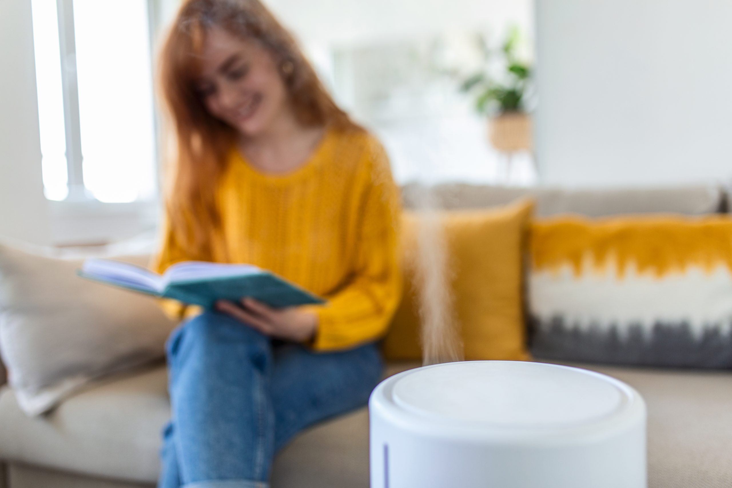 woman in background sitting on sofa with humidifer in forground in focus for banner for a post about why calgary air is so dry and solutions for huidity